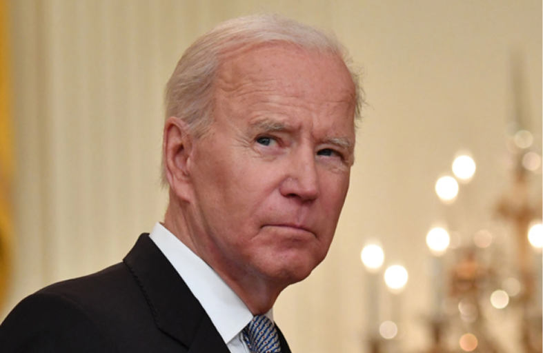 An elderly man in a suit looks to the side, standing indoors with chandeliers in the background.